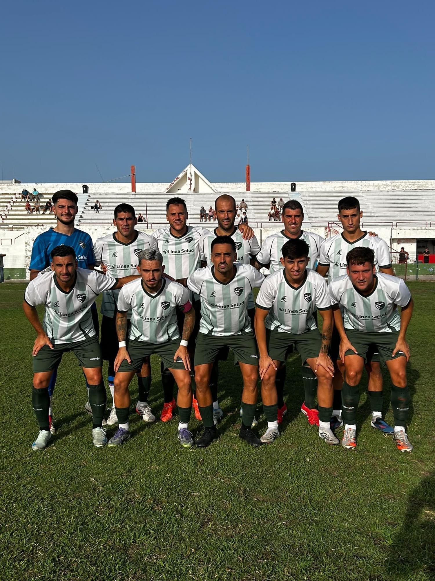Once jugadores de fútbol posan en dos filas sobre el césped de un campo, con gradas blancas al fondo.