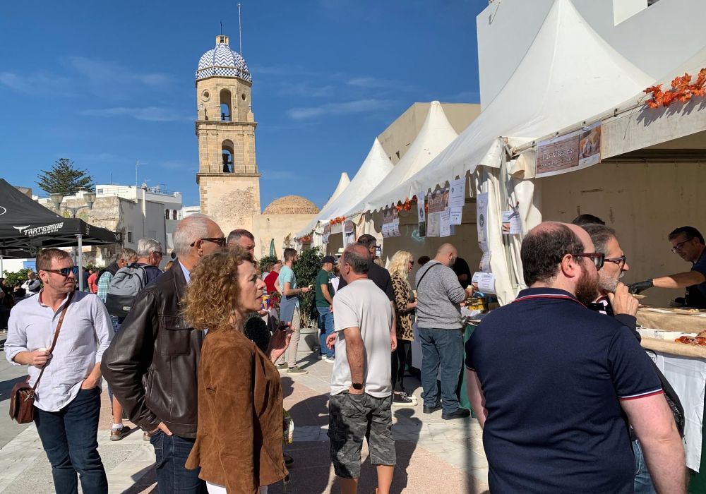 Multitud de personas junto a carpas blancas de un mercado al aire libre, con una torre de iglesia con cúpula al fondo.