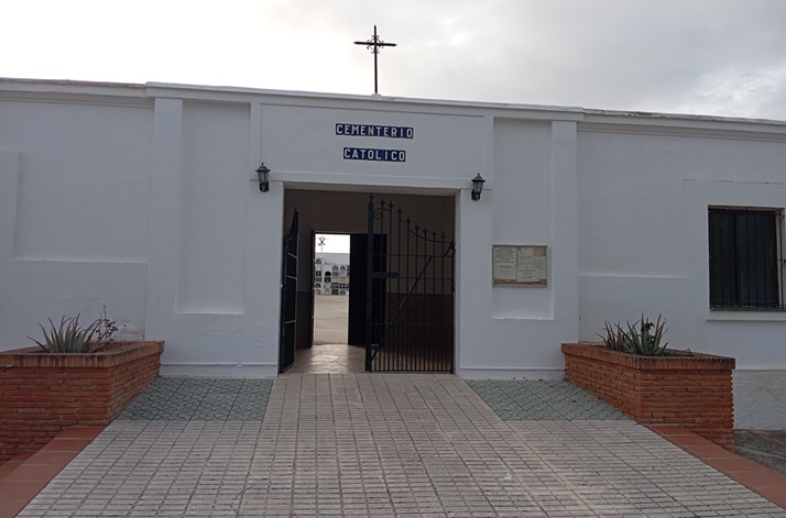 Fachada blanca de un cementerio católico con una cruz sobre la puerta de entrada abierta y dos jardineras de ladrillo.