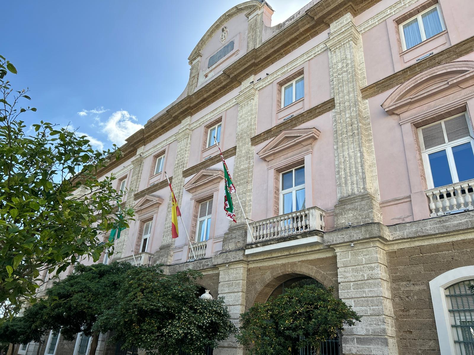 Fachada de un edificio institucional de color rosa con pilastras de piedra, balcones, banderas y árboles en primer plano.
