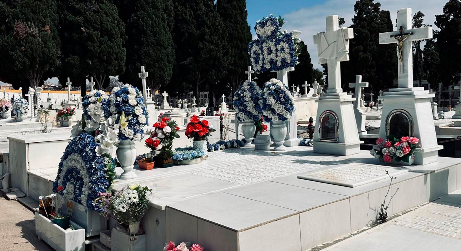 Tumbas blancas de mármol con cruces y coronas de flores azules, blancas y rojas en un cementerio con cipreses al fondo.