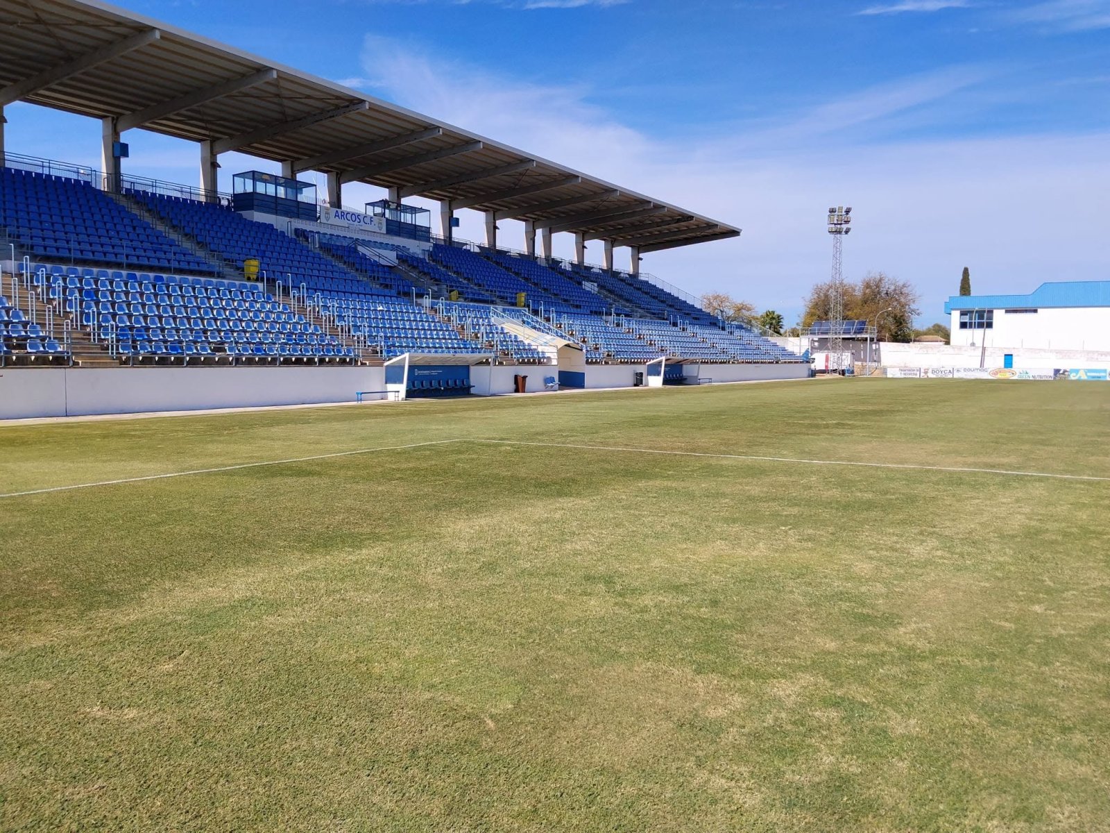 Campo de fútbol vacío con césped algo seco, gradas azules cubiertas y banquillos blancos bajo un cielo despejado.
