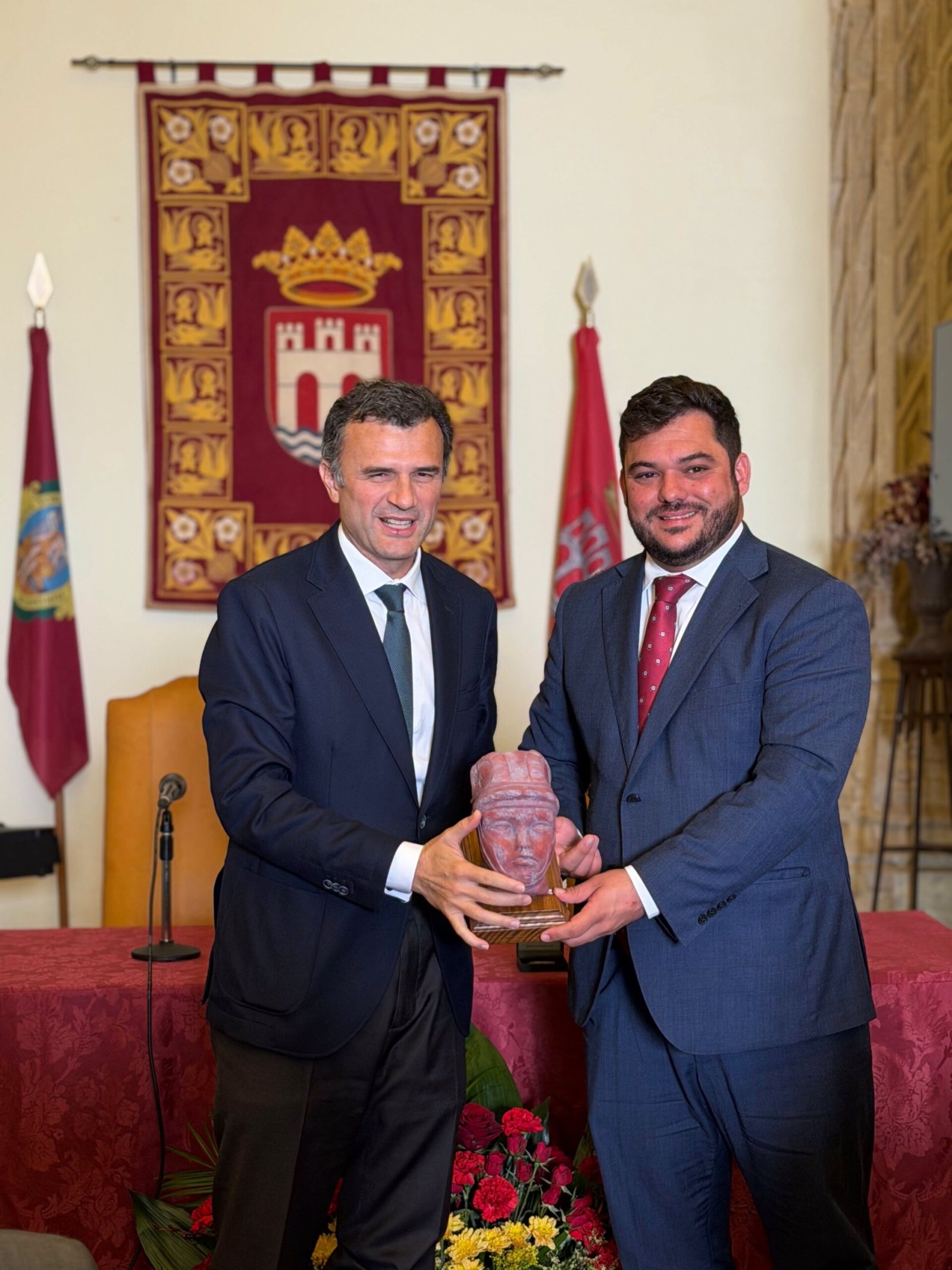 Dos hombres trajeados sostienen juntos una escultura con forma de cabeza sobre una base, ante un tapiz y banderas.
