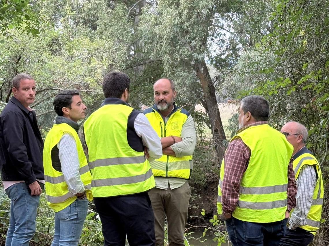 Un grupo de seis hombres con chalecos reflectantes amarillos conversa junto a una zona de vegetación y un cauce.