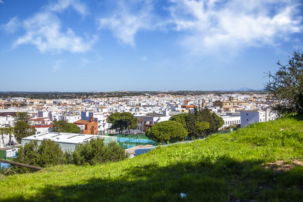 Vista panorámica de una ciudad de casas blancas y tejados, con árboles y una ladera verde en primer plano bajo un cielo azul con nubes.