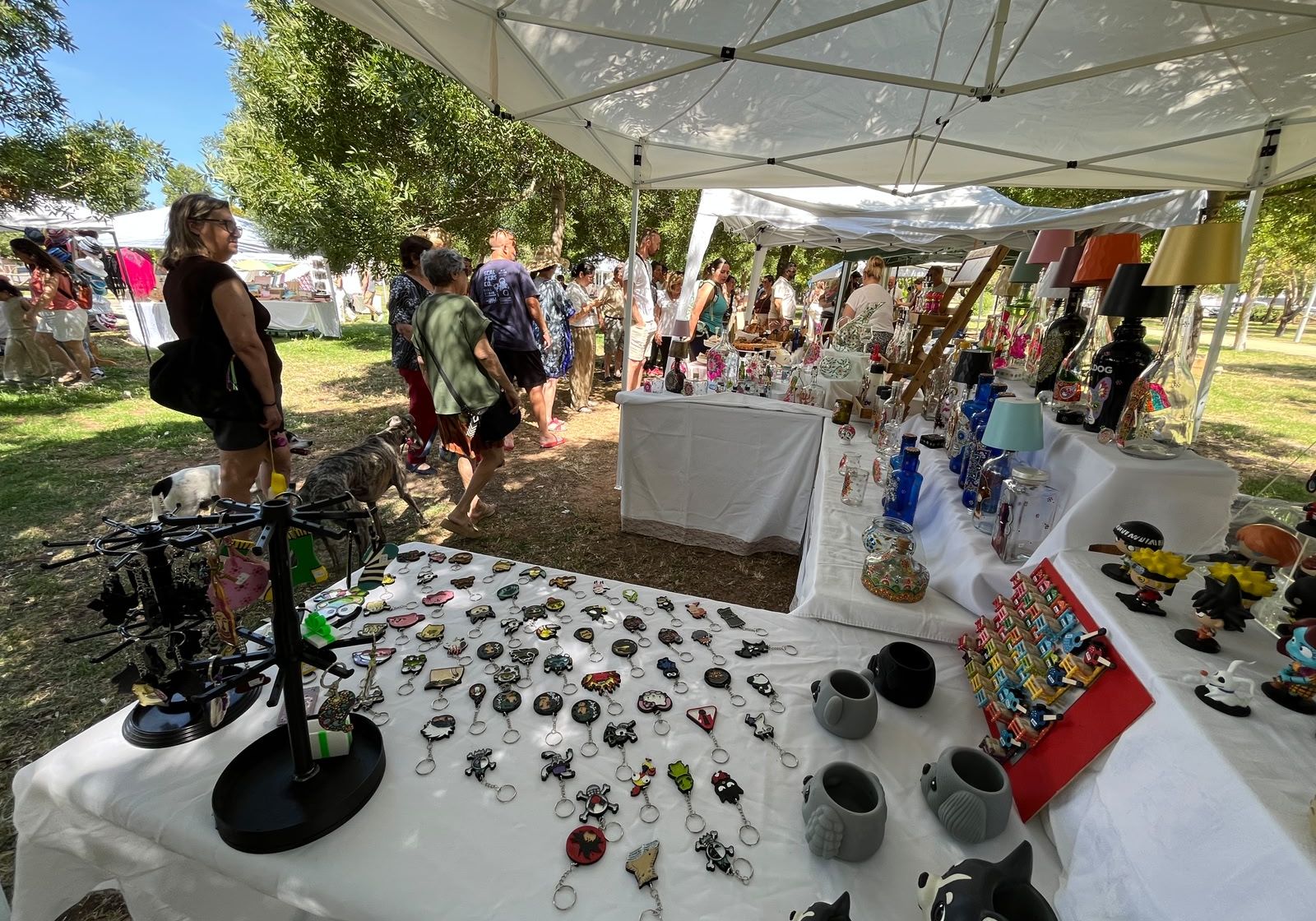 Puestos de artesanía bajo carpas blancas en un parque, con llaveros y figuras sobre una mesa y varias personas paseando alrededor.