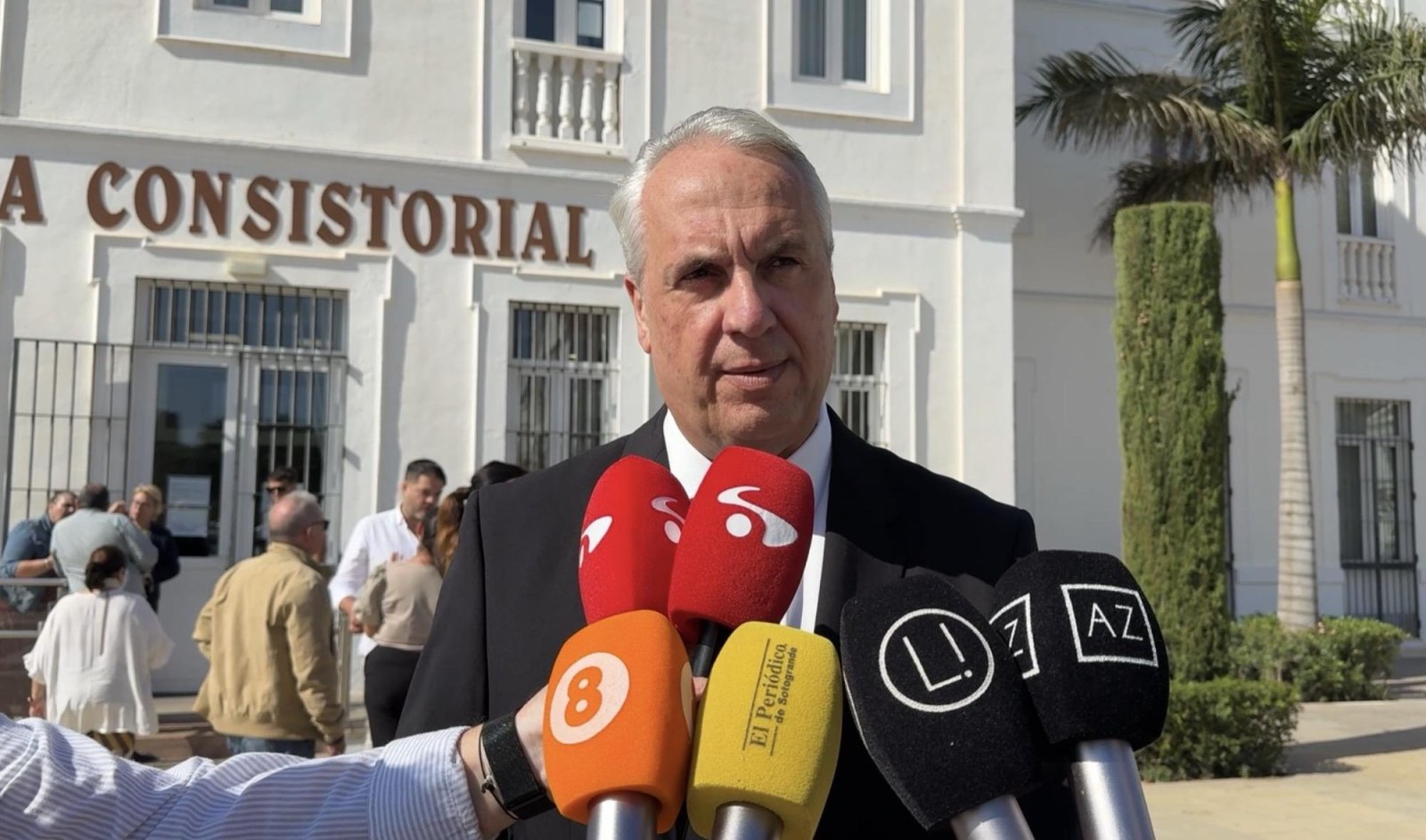 Un hombre mayor de traje y corbata habla ante varios micrófonos de medios, frente a un edificio blanco con el rótulo CONSISTORIAL.