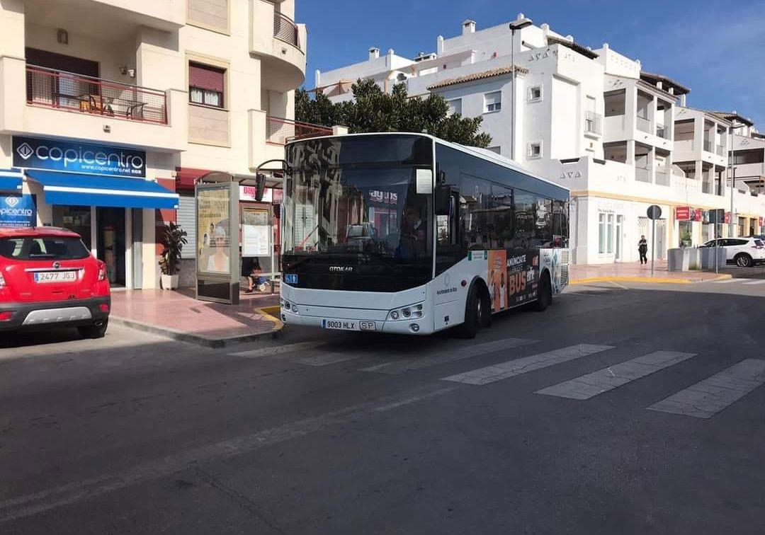 Un autobús blanco circula por una calle junto a una parada y edificios de varias plantas.