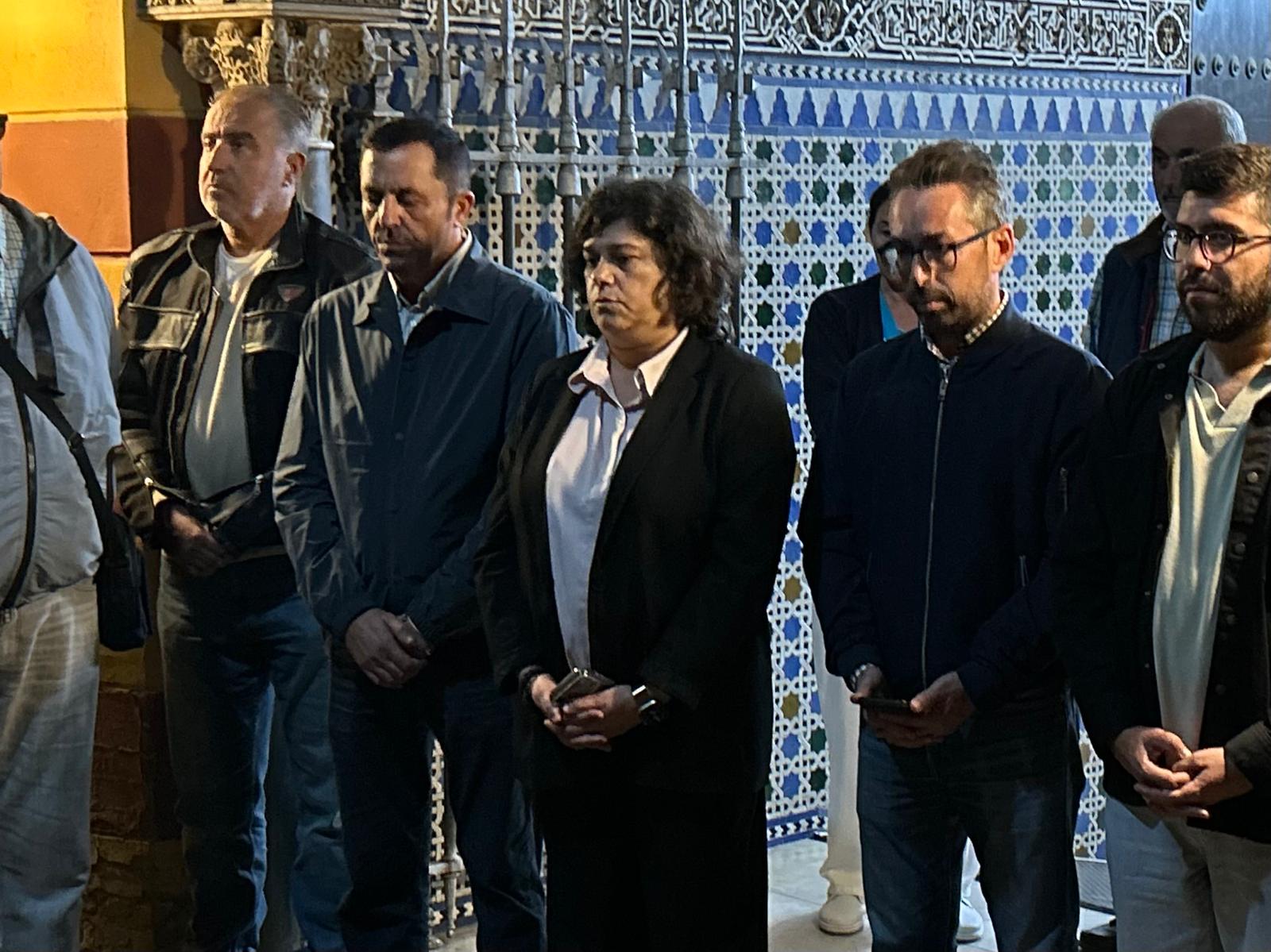 Un grupo de personas de pie, con gesto serio y las manos juntas, frente a una pared decorada con azulejos geométricos.