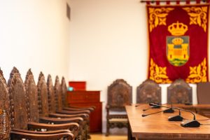 Sala de reuniones con una fila de sillas de madera tallada, una mesa con micrófonos y un tapiz rojo con escudo en la pared.