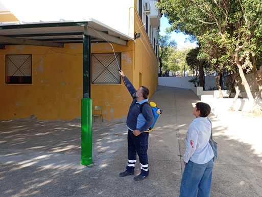 Un hombre con mochila pulverizadora aplica un chorro hacia la parte alta de un pilar verde junto a un edificio amarillo, observado por otra persona.