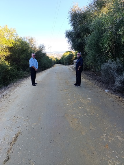 Dos hombres de pie sobre un camino de tierra flanqueado por vegetación, con campo al fondo.