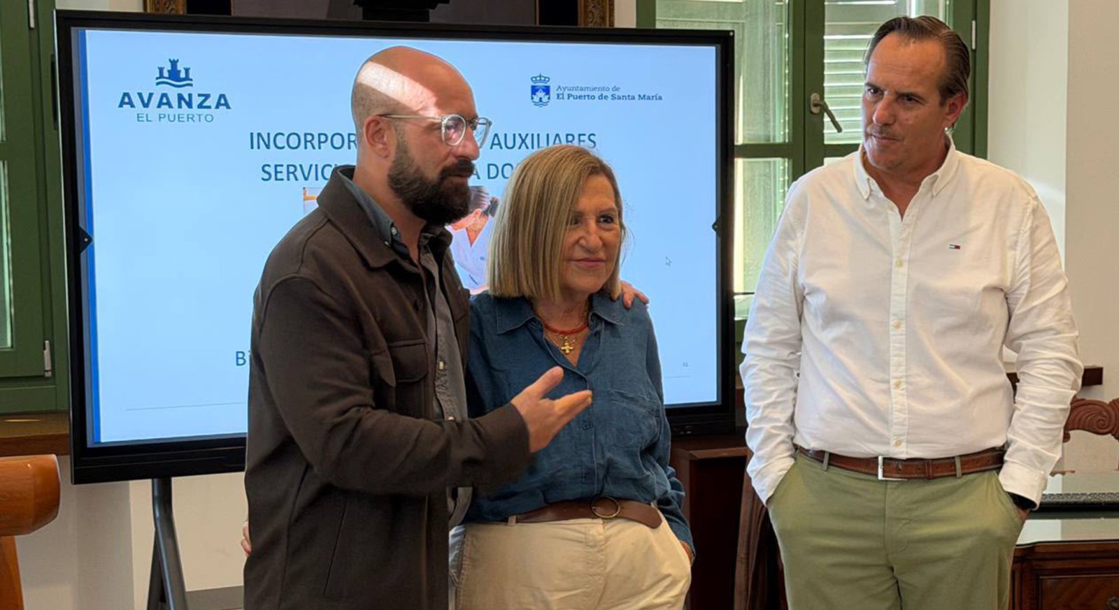 Tres personas de pie ante una pantalla grande que muestra el texto AVANZA EL PUERTO y AUXILIARES.