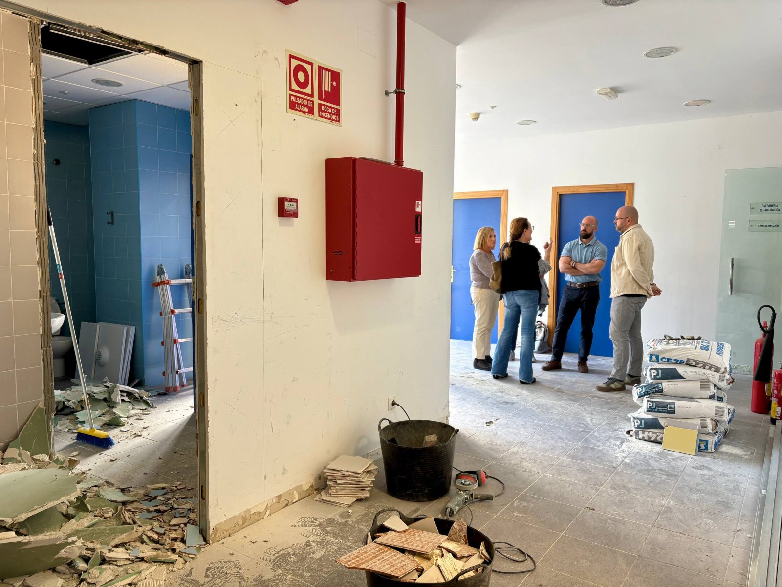 Interior en obras con escombros, cubos, sacos y cuatro personas conversando junto a puertas azules.
