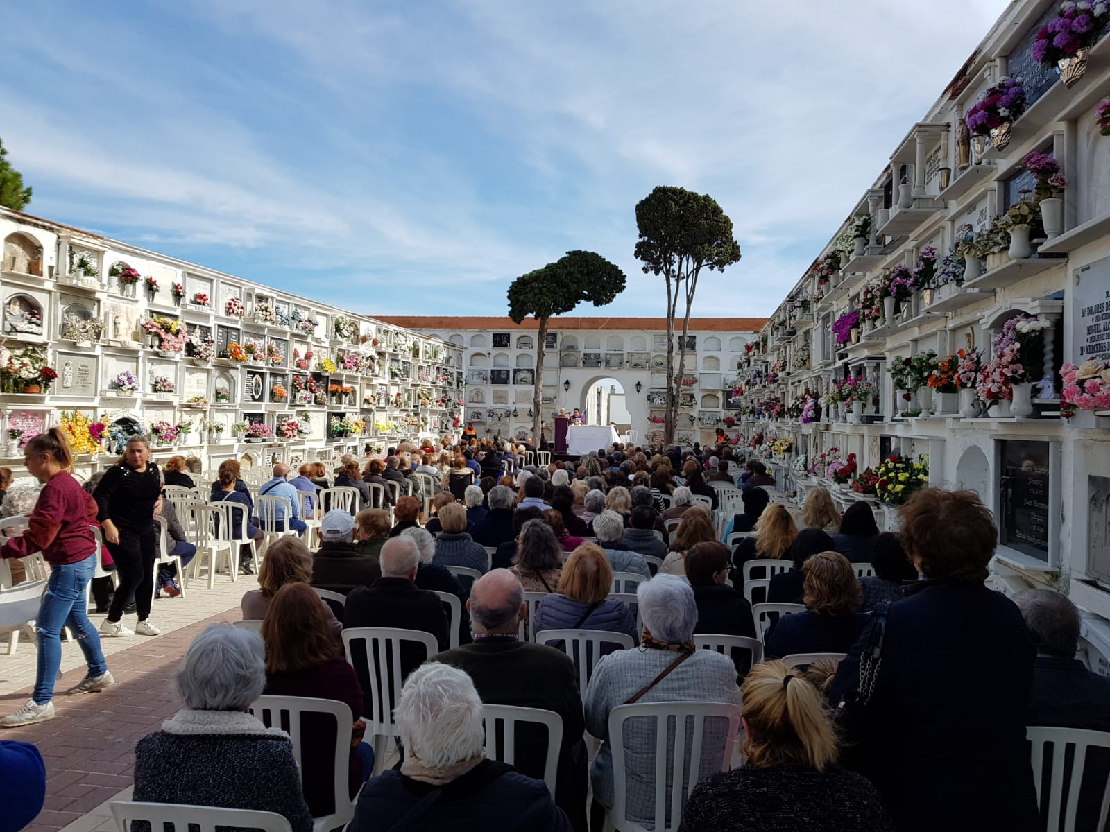 Multitud sentada en sillas blancas en el pasillo central de un cementerio con nichos florales a ambos lados.