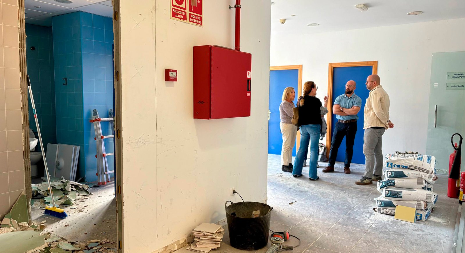 Pasillo en obras con escombros, escalera, sacos y cuatro personas conversando junto a puertas azules.