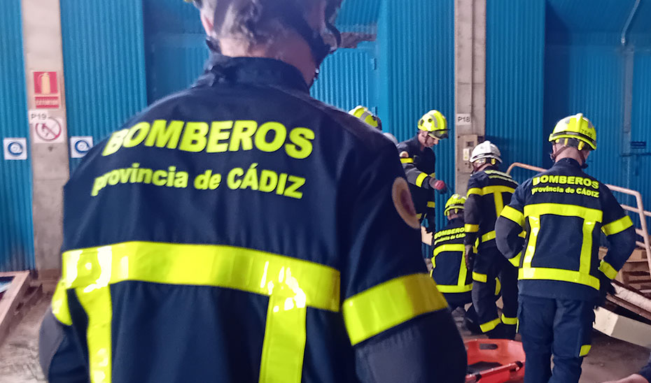 Varios bomberos con trajes azul marino y franjas amarillas, con la inscripción Bomberos provincia de Cádiz, reunidos en el interior de una nave industrial.