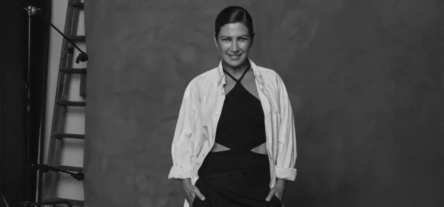 Mujer sonriente con camisa blanca abierta sobre top negro, posando frente a un fondo liso en un estudio.