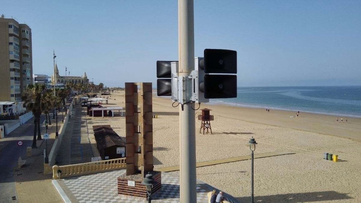 Una playa amplia con paseo, palmeras y edificios al fondo, y en primer plano un poste con altavoces negros junto a una torre de ladrillo.