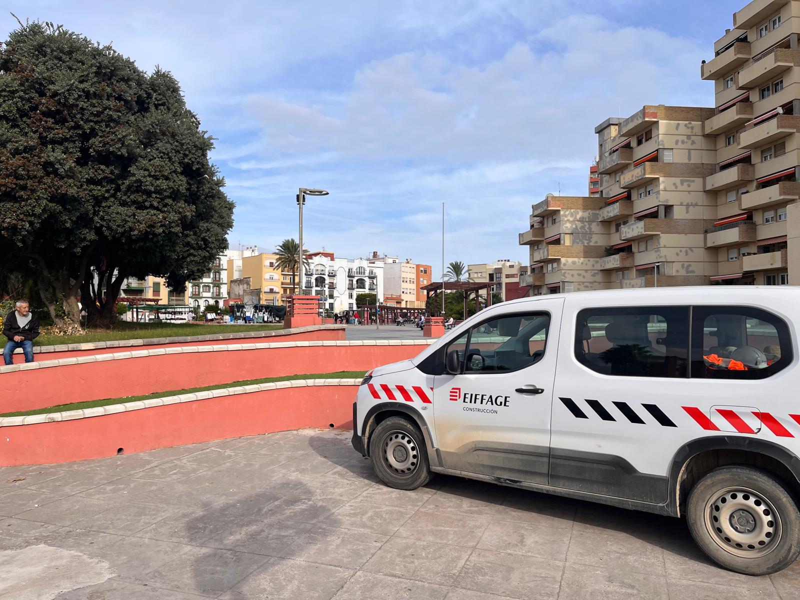 Una furgoneta blanca de construcción aparcada junto a un muro bajo rojo, con un hombre sentado y edificios al fondo.