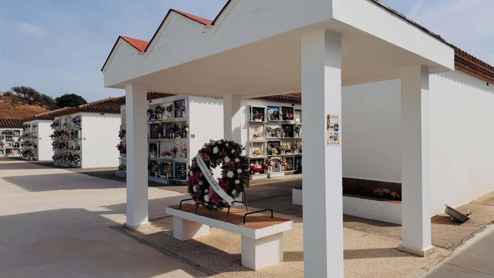 Patio de un cementerio con nichos blancos, una corona de flores sobre un banco y una pérgola de pilares blancos.