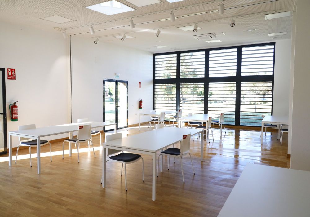 Sala amplia y vacía con mesas blancas, sillas de asiento oscuro, suelo de madera y grandes ventanales con lamas.