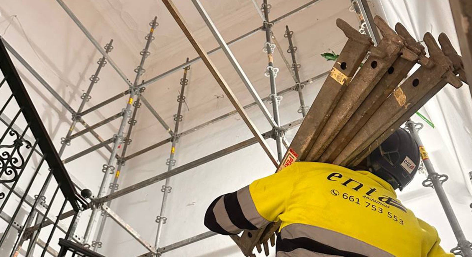 Un operario con camiseta amarilla y casco carga barras metálicas de andamio bajo una estructura de andamiaje y un techo blanco.