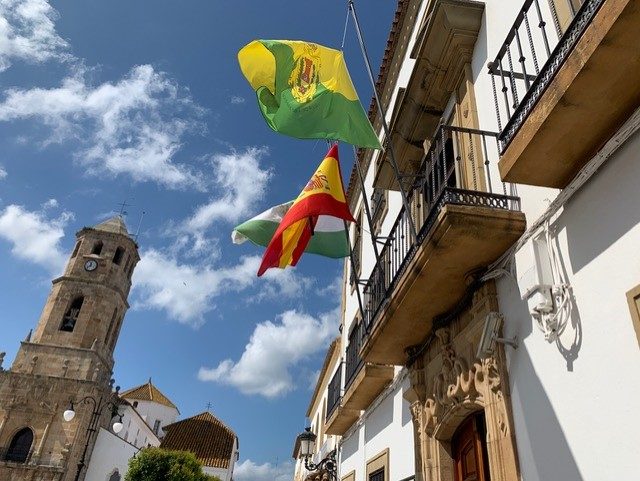 Una torre de iglesia con reloj a la izquierda y, a la derecha, una fachada blanca con balcones y banderas ondeando bajo un cielo azul con nubes.