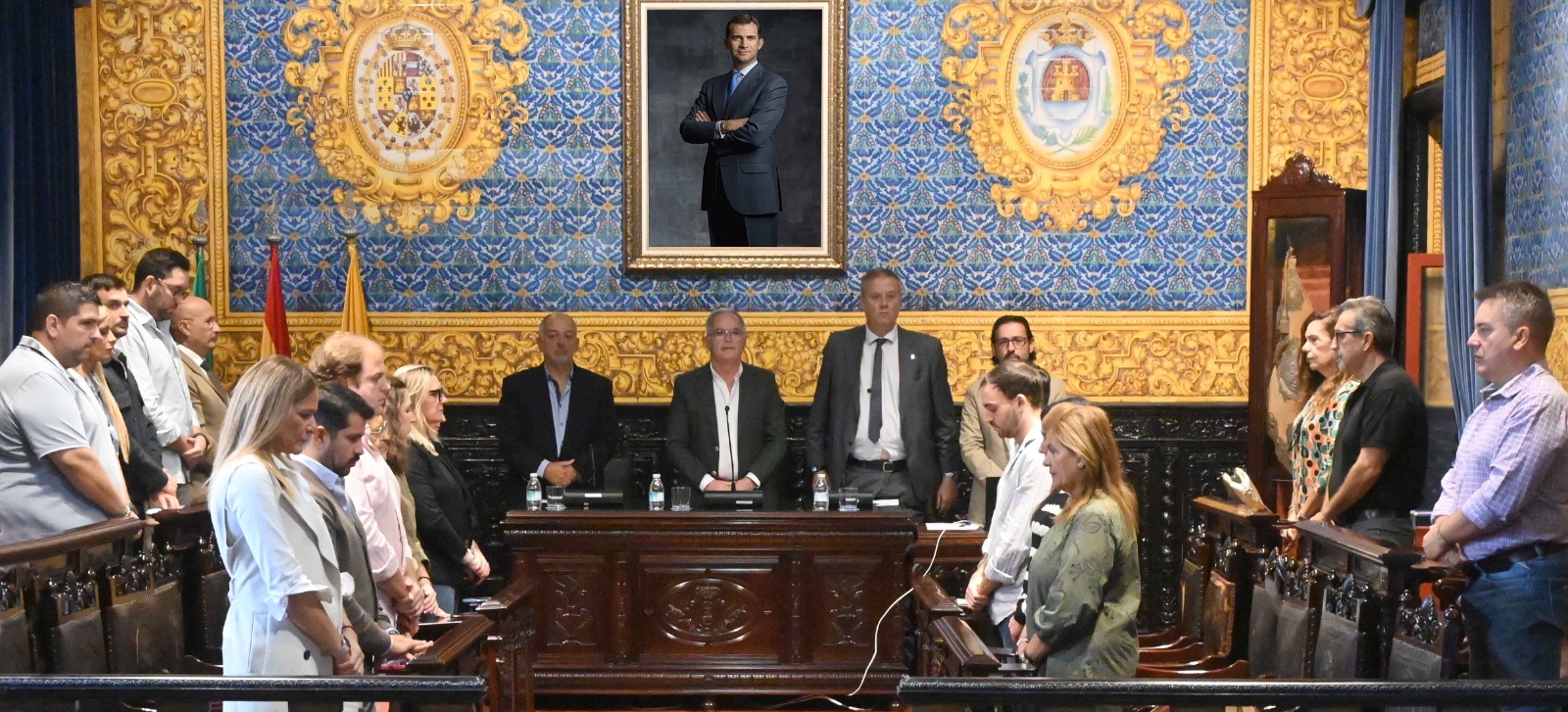 Un grupo de personas de pie en una sala con paredes decoradas en azul y dorado y un retrato enmarcado al fondo.