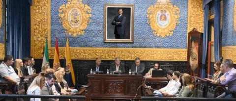 Personas sentadas en bancos de madera ante una mesa presidencial en una sala con paredes azules y doradas, escudos y un retrato.