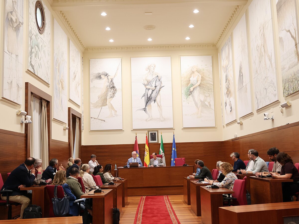 Sala de reuniones con público sentado en bancos de madera y una mesa presidencial con banderas y grandes pinturas murales.