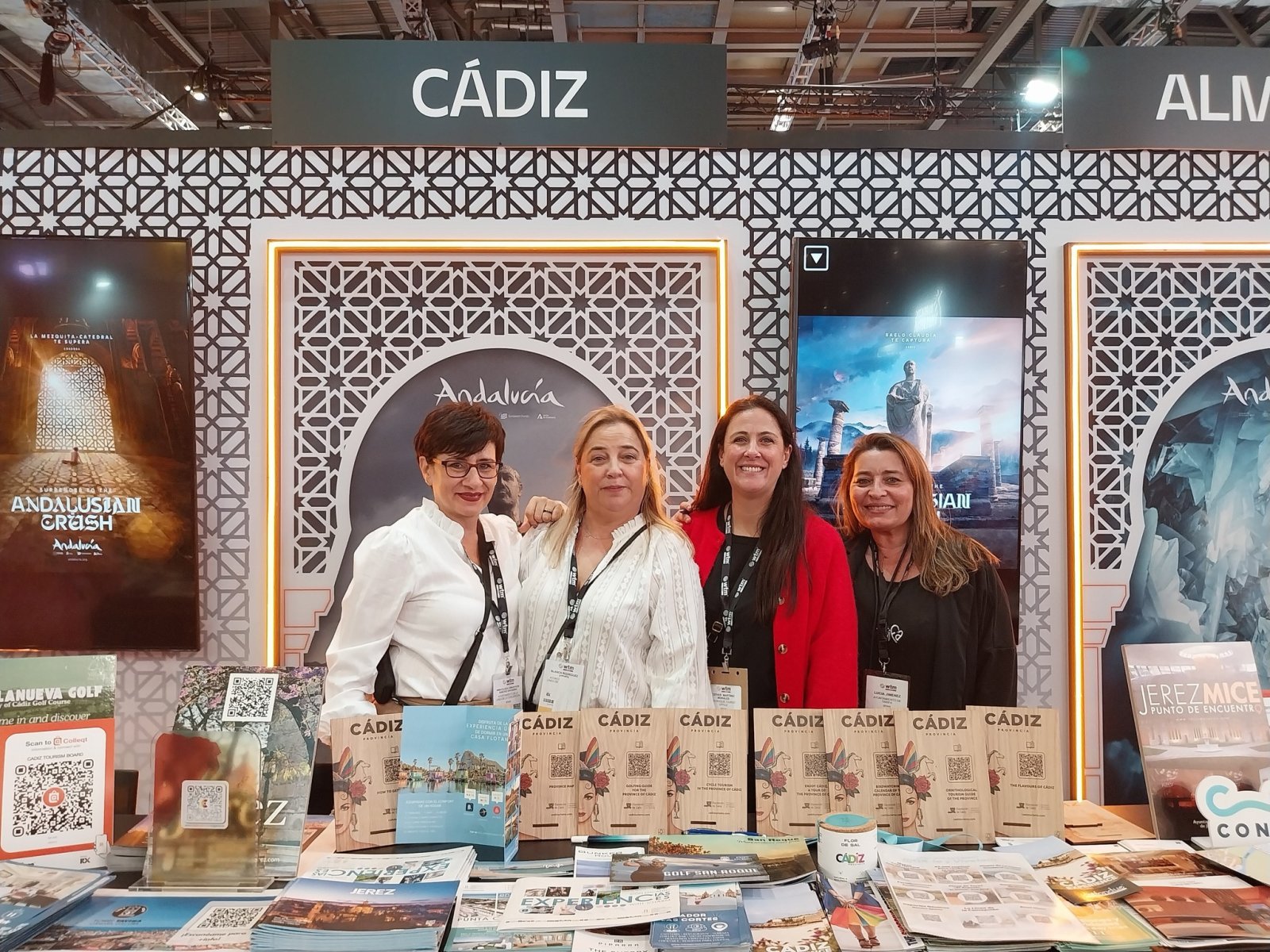 Cuatro mujeres posan sonrientes tras un mostrador con folletos y carteles de Cádiz y Andalucía en un stand de feria.
