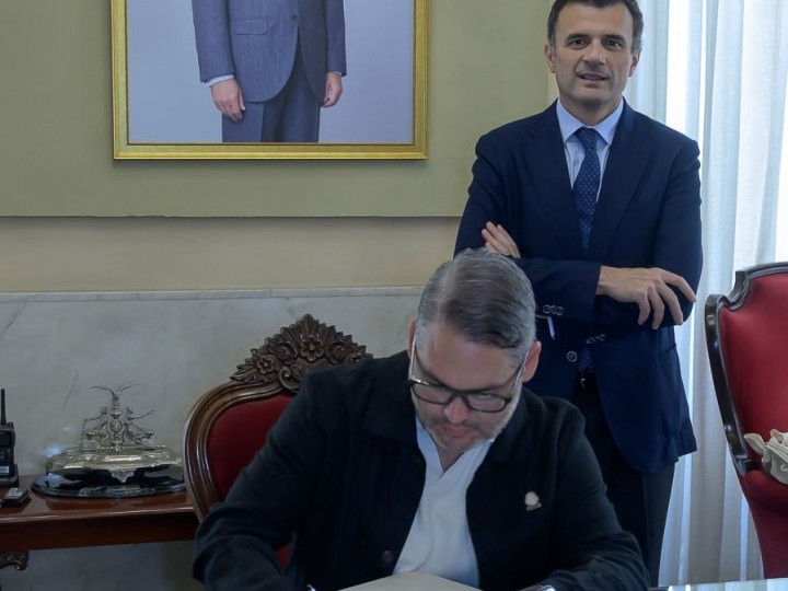 Dos hombres trajeados en una sala: uno sentado firmando y otro de pie con los brazos cruzados, ante un retrato enmarcado.